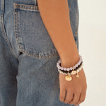 A close-up of a person's wrist wearing a stack of beaded bracelets, including light purple, dark brown, and gold charms, paired with blue denim jeans against a plain white background.