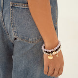 A close-up of a person's wrist wearing a stack of beaded bracelets, including light purple, dark brown, and gold charms, paired with blue denim jeans against a plain white background.