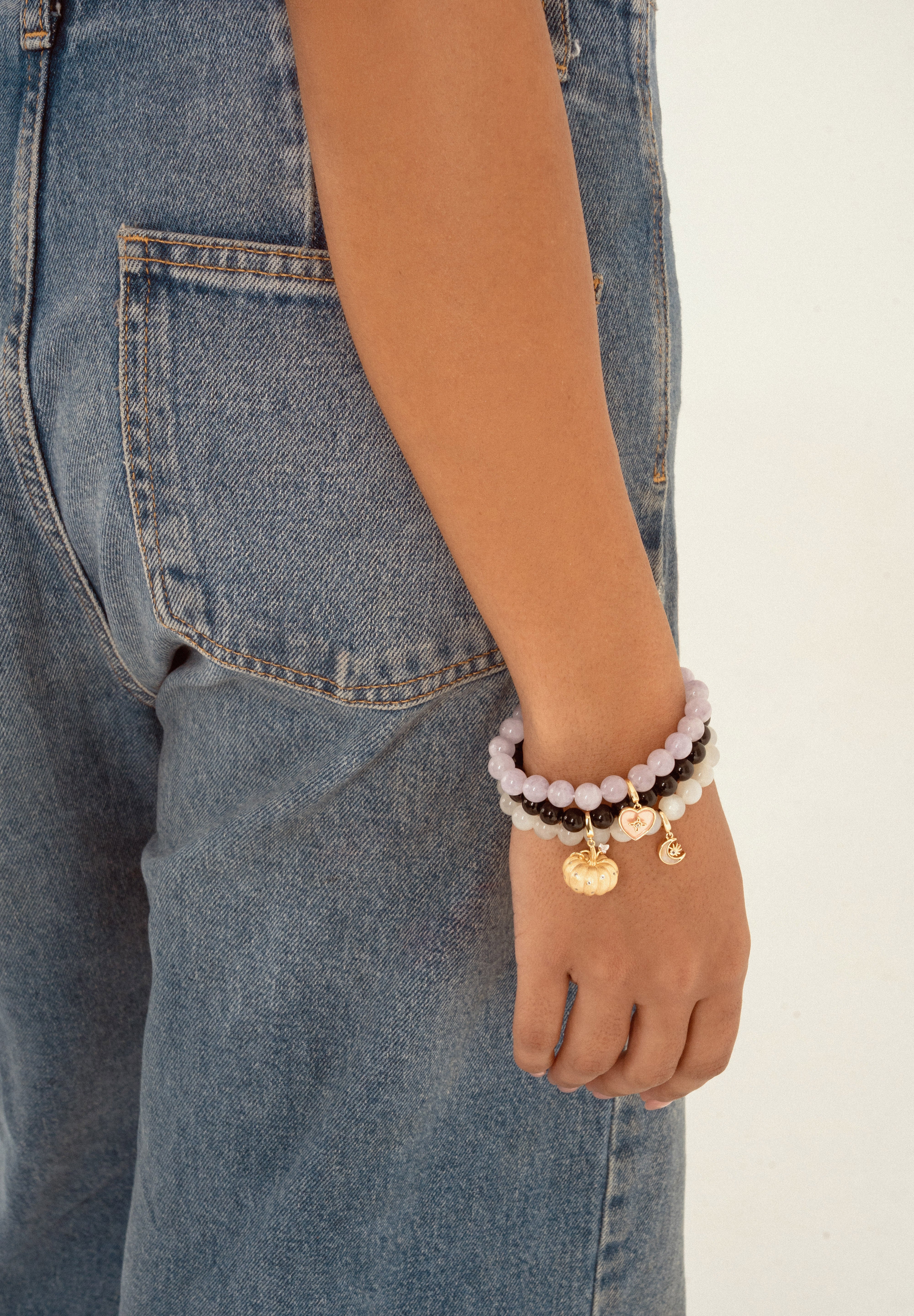 A close-up of a person's wrist wearing a stack of beaded bracelets, including light purple, dark brown, and gold charms, paired with blue denim jeans against a plain white background.