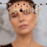 A close-up of a model with short brown hair holding up a delicate gold chain across her forehead, adorned with multiple small charms against a soft gray background.