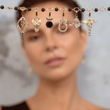 A close-up of a model with short brown hair holding up a delicate gold chain across her forehead, adorned with multiple small charms against a soft gray background.