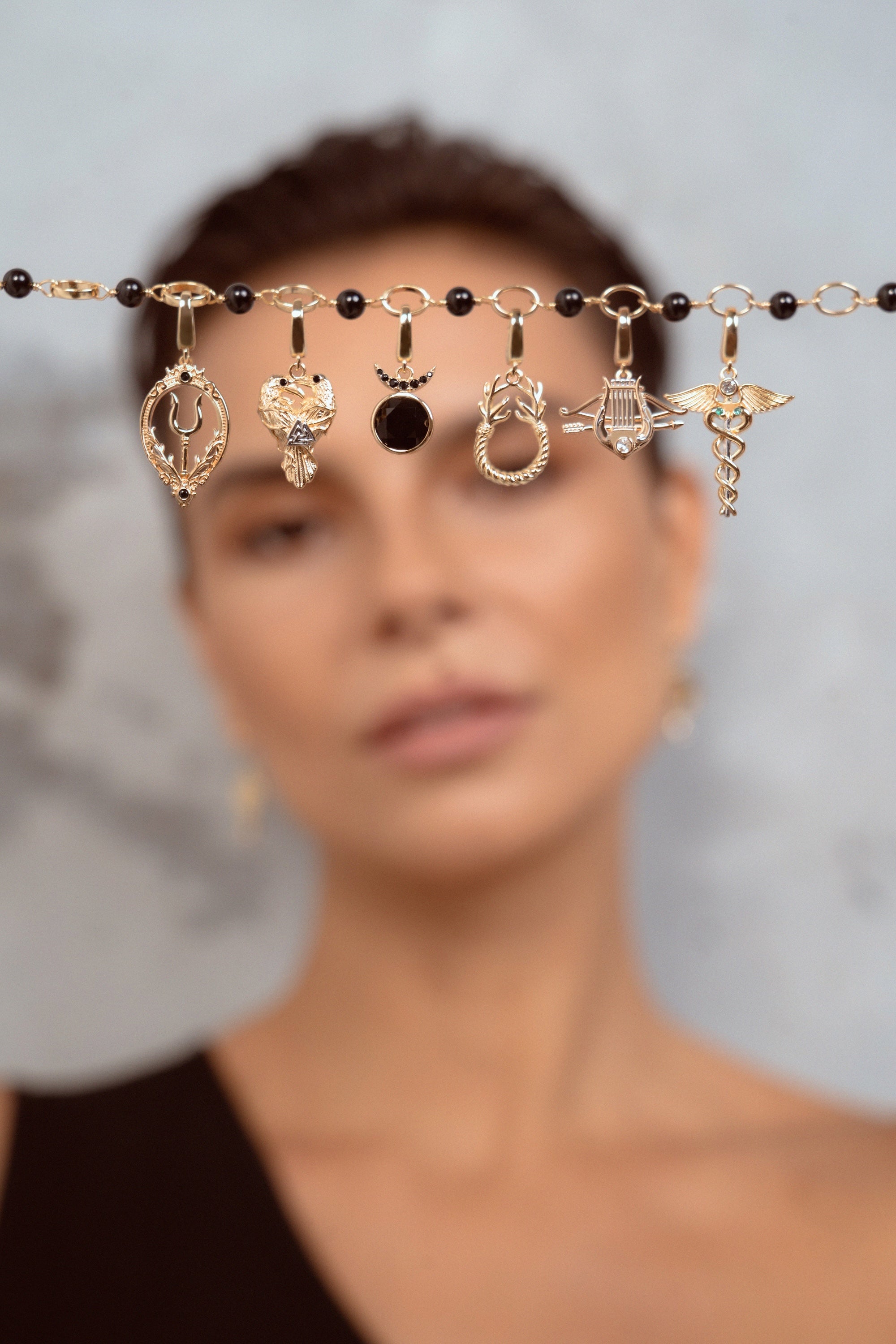 A close-up of a model with short brown hair holding up a delicate gold chain across her forehead, adorned with multiple small charms against a soft gray background.