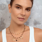 A close-up of a woman wearing a double-strand beaded necklace with gold charms, over a white ribbed tank top against a textured gray background.