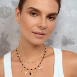 A close-up of a woman wearing a double-strand beaded necklace with gold charms, over a white ribbed tank top against a textured gray background.