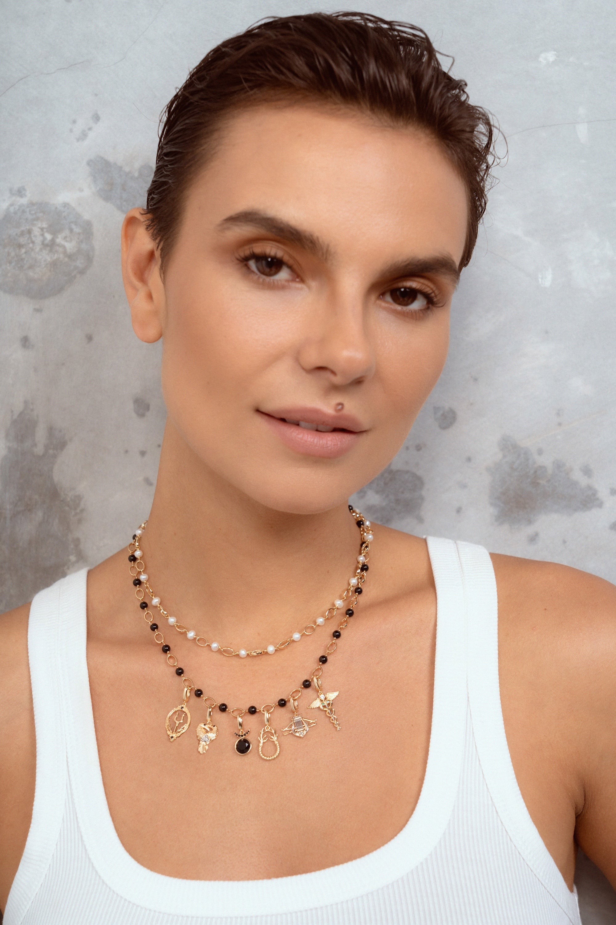 A close-up of a woman wearing a double-strand beaded necklace with gold charms, over a white ribbed tank top against a textured gray background.