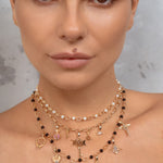 A close-up of a model wearing a multi-layered gold necklace with black beads, white pearls, and various celestial and zodiac-themed charms, including a key, moon, and stars.