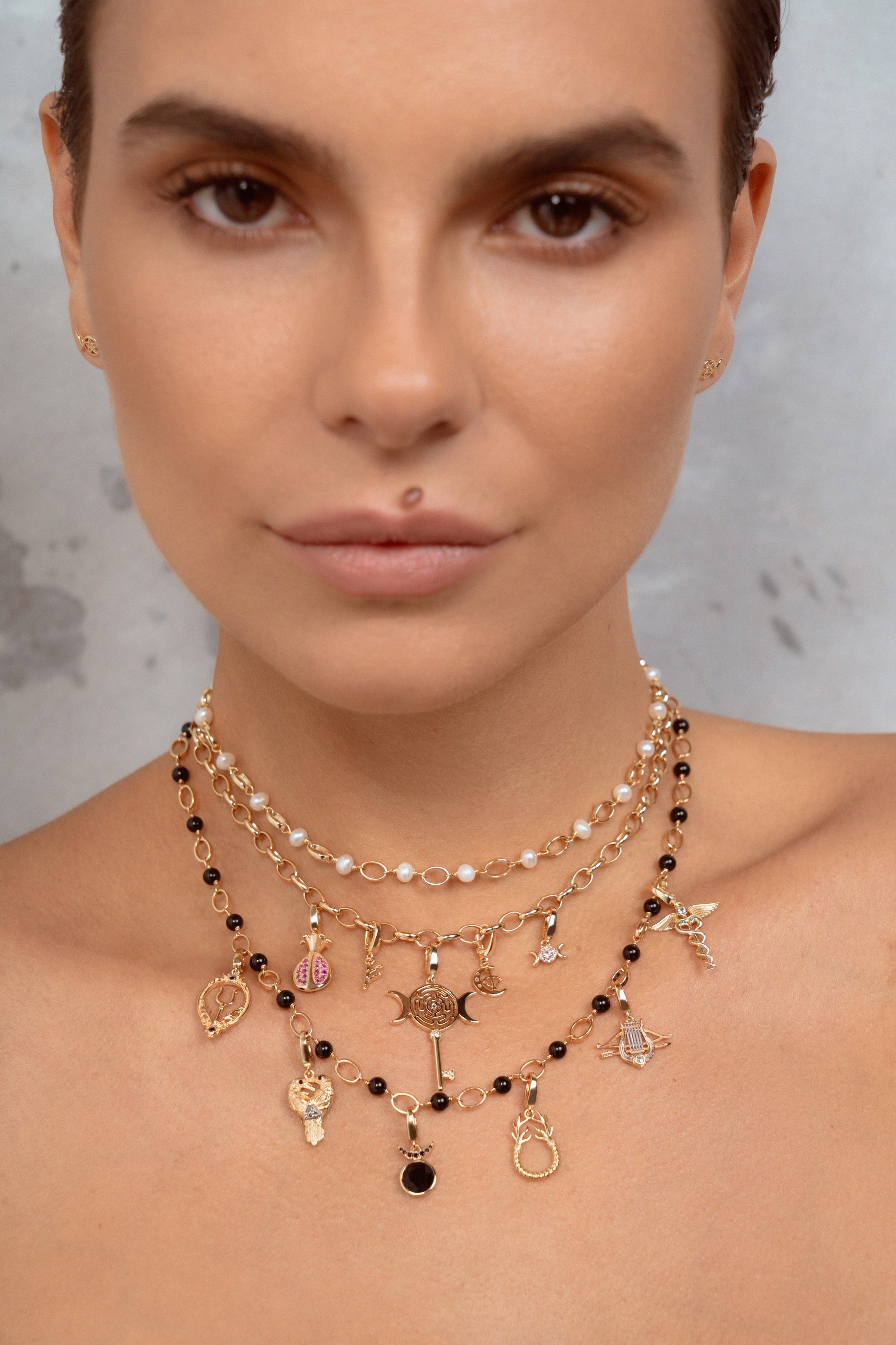 A close-up of a model wearing a multi-layered gold necklace with black beads, white pearls, and various celestial and zodiac-themed charms, including a key, moon, and stars.