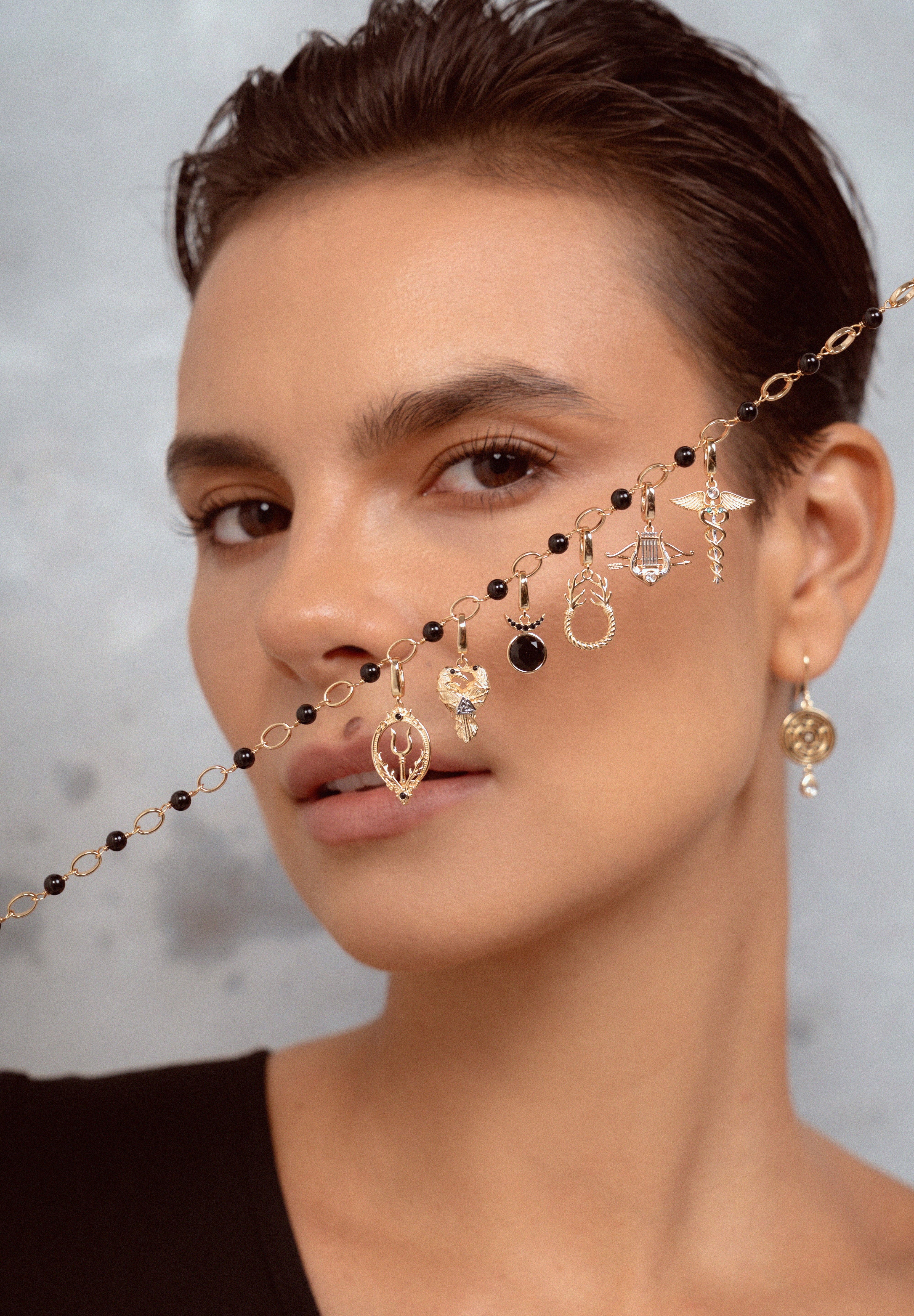 A close-up of a model wearing a delicate gold chain necklace with multiple dangling charms, including black beads, clear crystal pendants, and small gold elements. The model is shown from the chest up against a textured gray background.