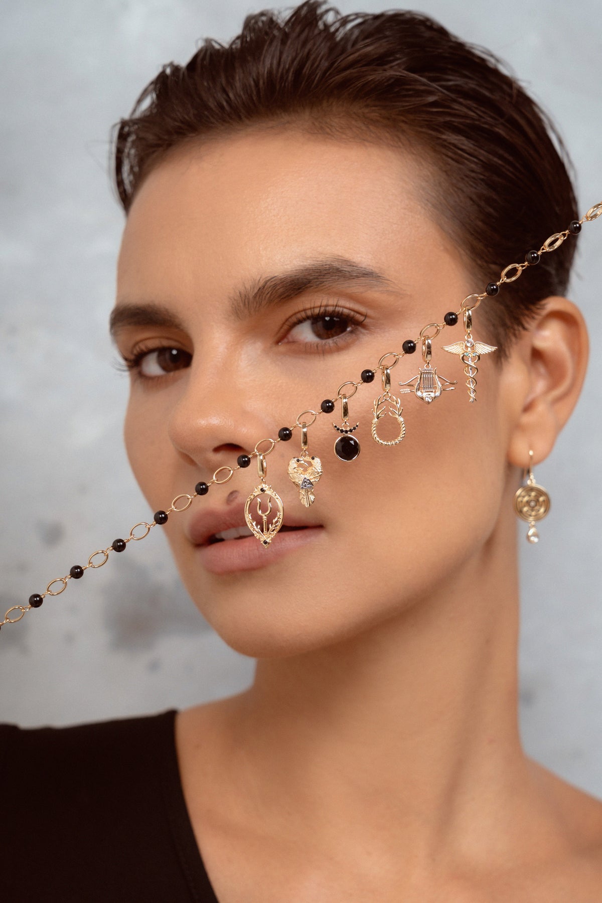 A close-up of a model wearing a delicate gold chain necklace with multiple dangling charms, including black beads, clear crystal pendants, and small gold elements. The model is shown from the chest up against a textured gray background.