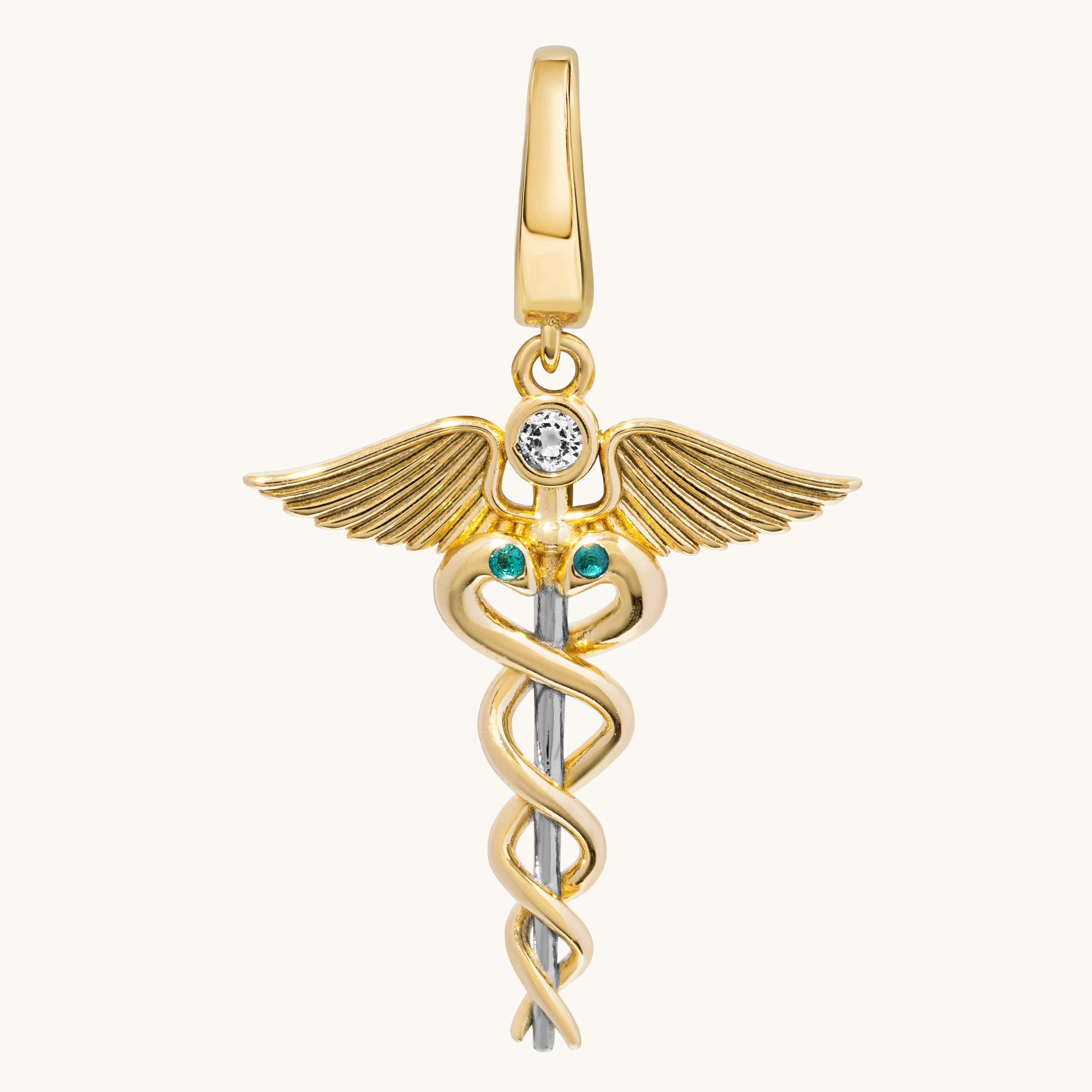 A gold medical caduceus pendant charm featuring a gold winged figure with a clear round gemstone and two small green emerald accents on the staff, shown on a light cream background.