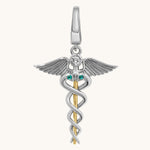 A two-tone silver and gold caduceus pendant charm featuring a silver winged figure with a clear round gemstone and two small green emerald accents, suspended from a gold staff on a light cream background.