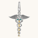 A two-tone silver and gold caduceus pendant charm featuring a silver winged figure with a clear round gemstone and two small green emerald accents, suspended from a gold staff on a light cream background.