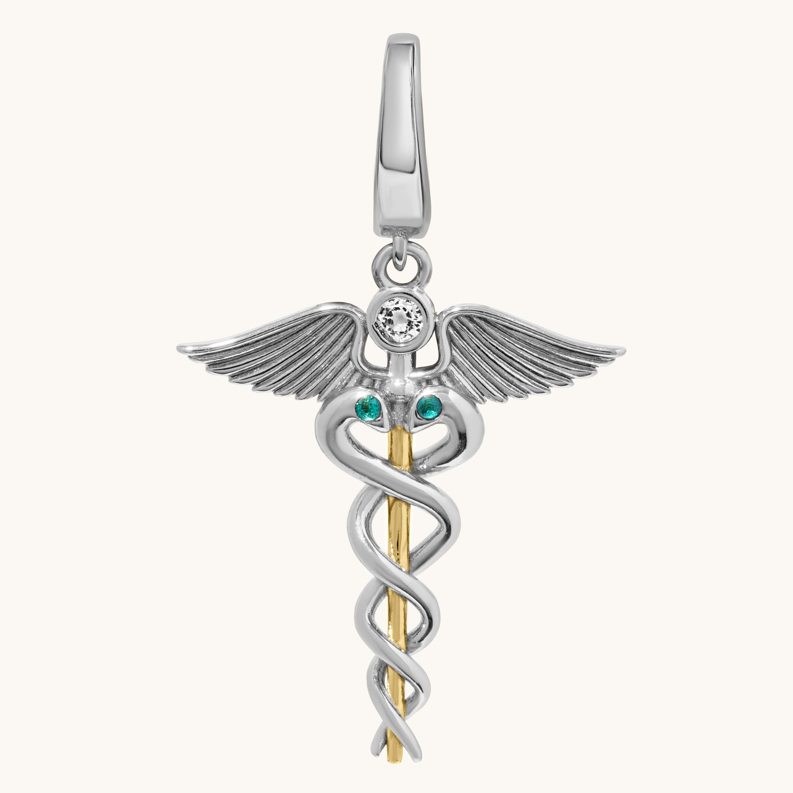 A two-tone silver and gold caduceus pendant charm featuring a silver winged figure with a clear round gemstone and two small green emerald accents, suspended from a gold staff on a light cream background.
