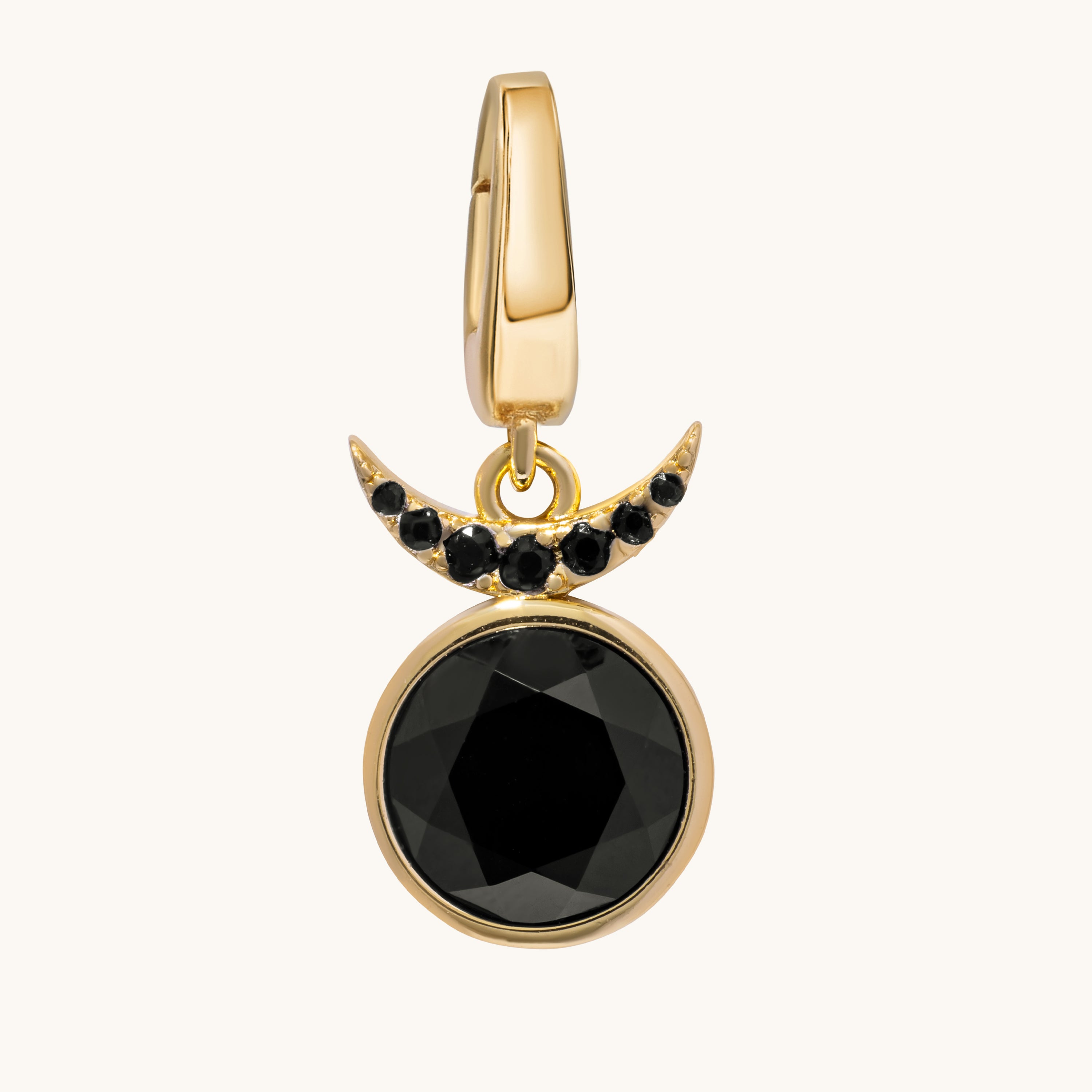 A gold crescent moon and large faceted black gemstone pendant charm, accented with small black stones, shown on a light cream background.