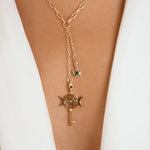 A gold-toned Y-necklace with a paperclip chain, a key and crescent moon pendant, and a small green gemstone star charm, shown worn on a model's neck over a white blazer.
