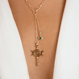 A gold-toned Y-necklace with a paperclip chain, a key and crescent moon pendant, and a small green gemstone star charm, shown worn on a model's neck over a white blazer.