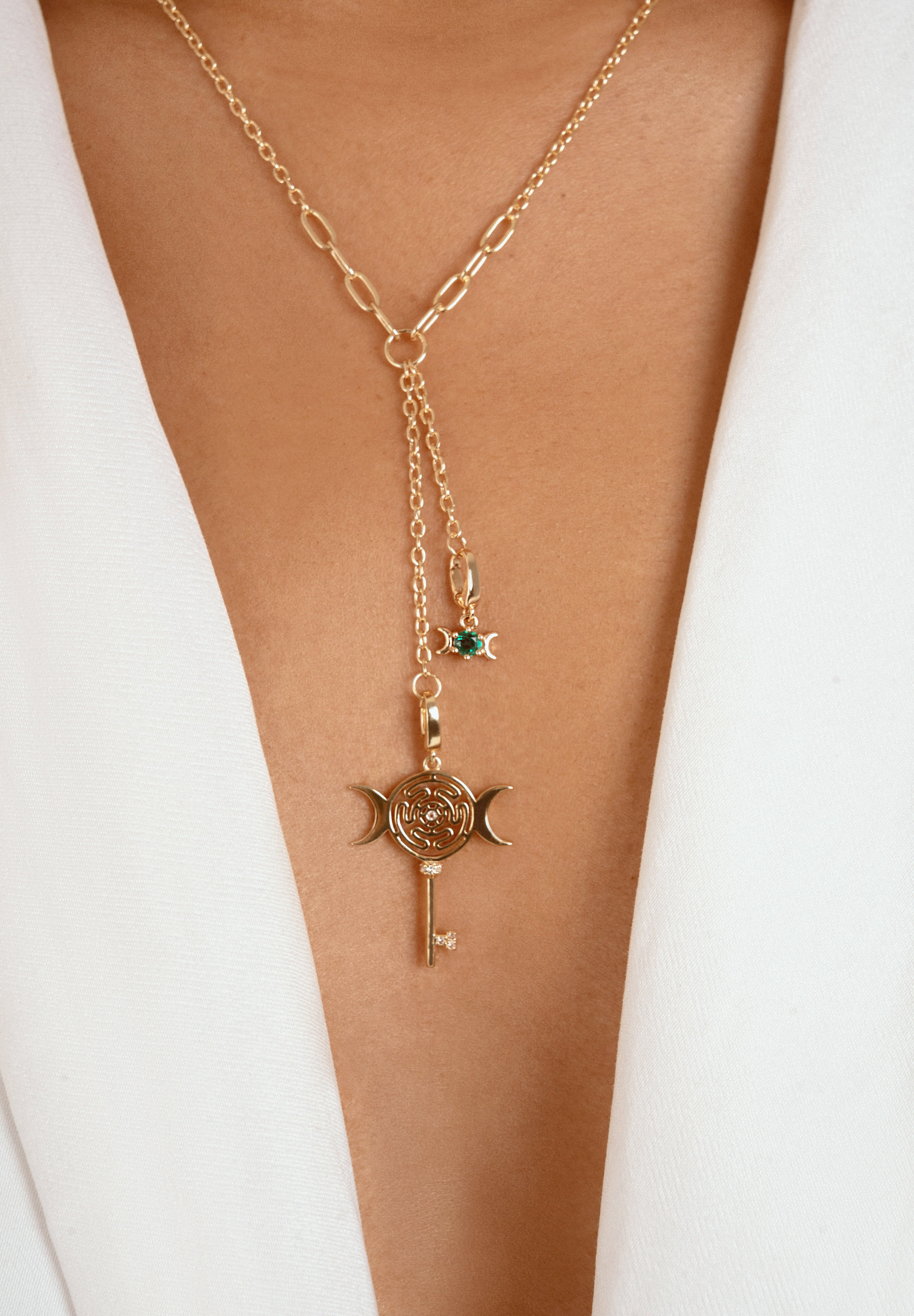 A gold-toned Y-necklace with a paperclip chain, a key and crescent moon pendant, and a small green gemstone star charm, shown worn on a model's neck over a white blazer.