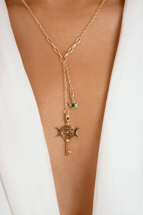 A gold-toned Y-necklace with a paperclip chain, a key and crescent moon pendant, and a small green gemstone star charm, shown worn on a model's neck over a white blazer.