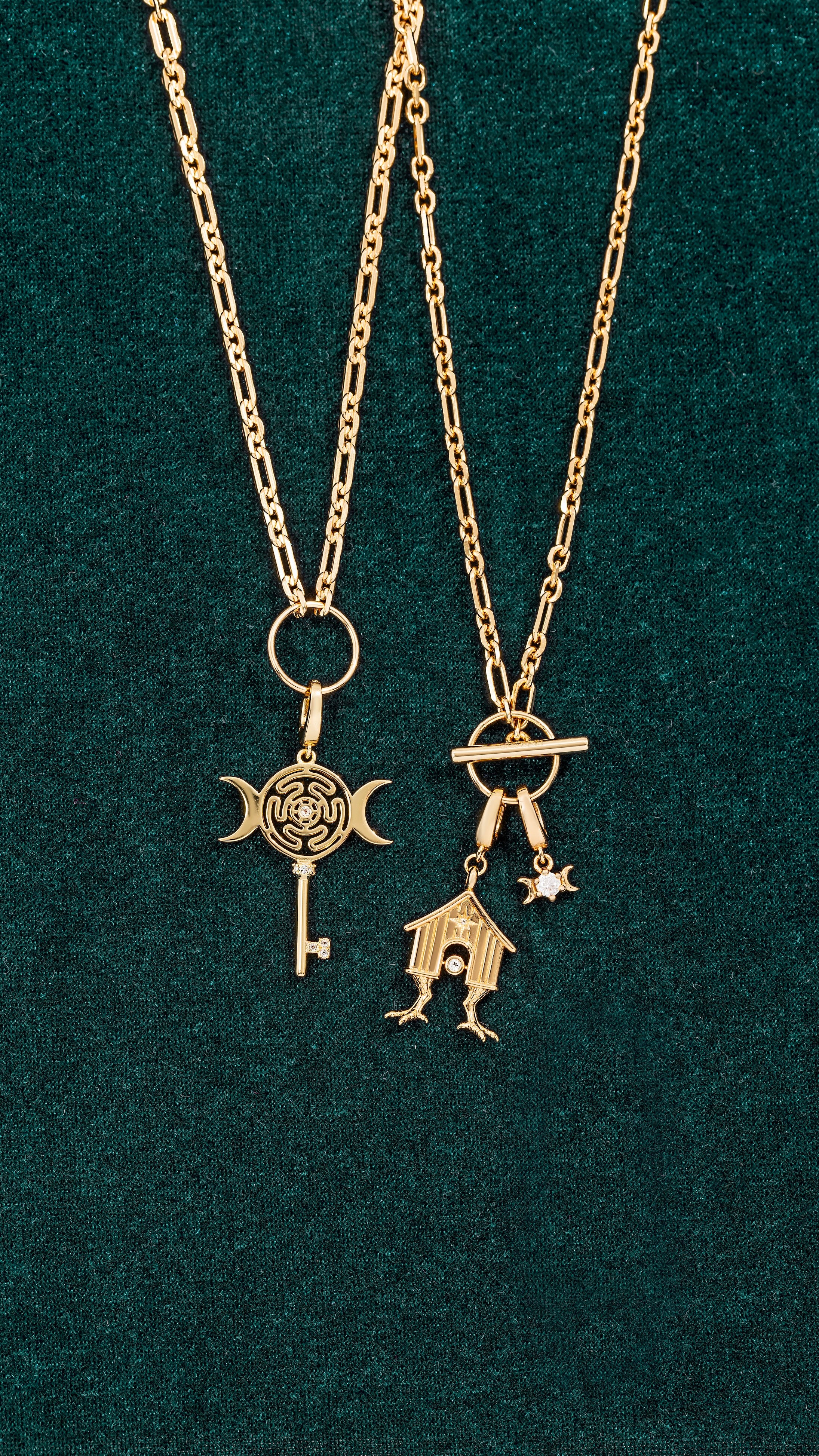 Two gold necklaces on dark green fabric: a reversible charm necklace with key and crescent moons, and a Magick Keeper Reversible Charm Necklace featuring a house charm, legs, stars, and crescent moon accents.
