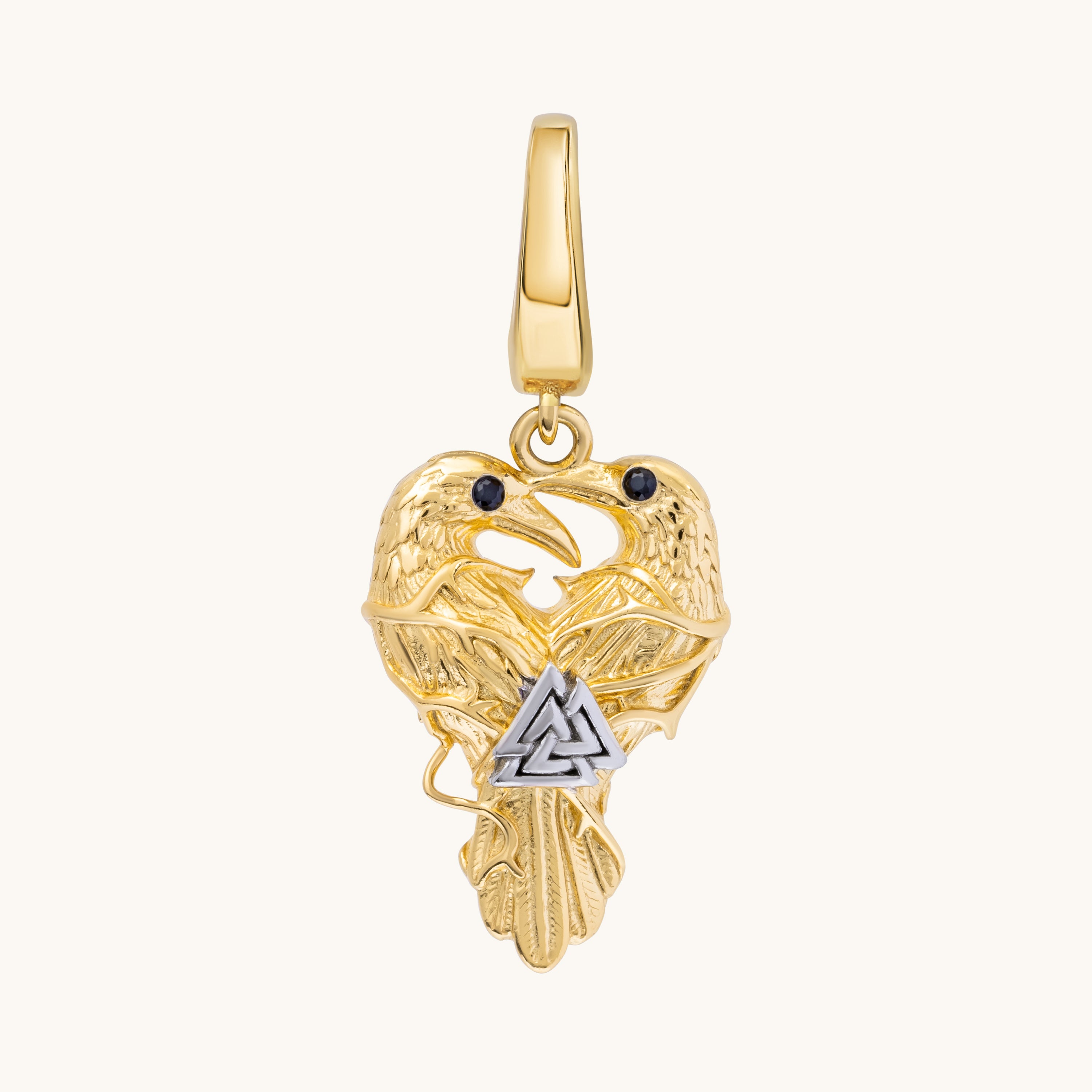 A gold pendant featuring two ravens facing each other with a silver-toned triangular knot motif at their intersection, set against a plain off-white background.