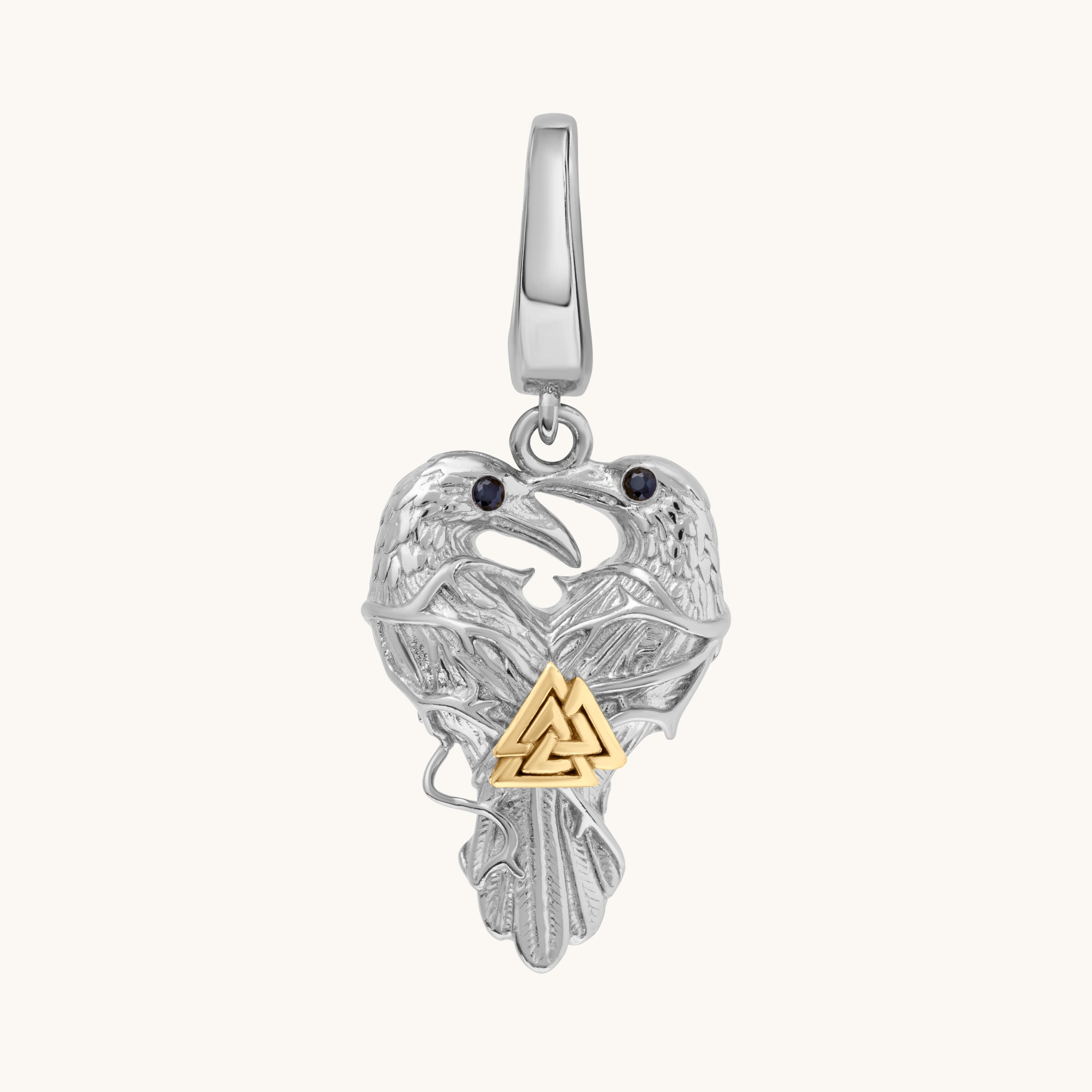 A sterling silver raven pendant with a gold Celtic triangle knot at the center and small black stone eyes, shown on a light cream background.