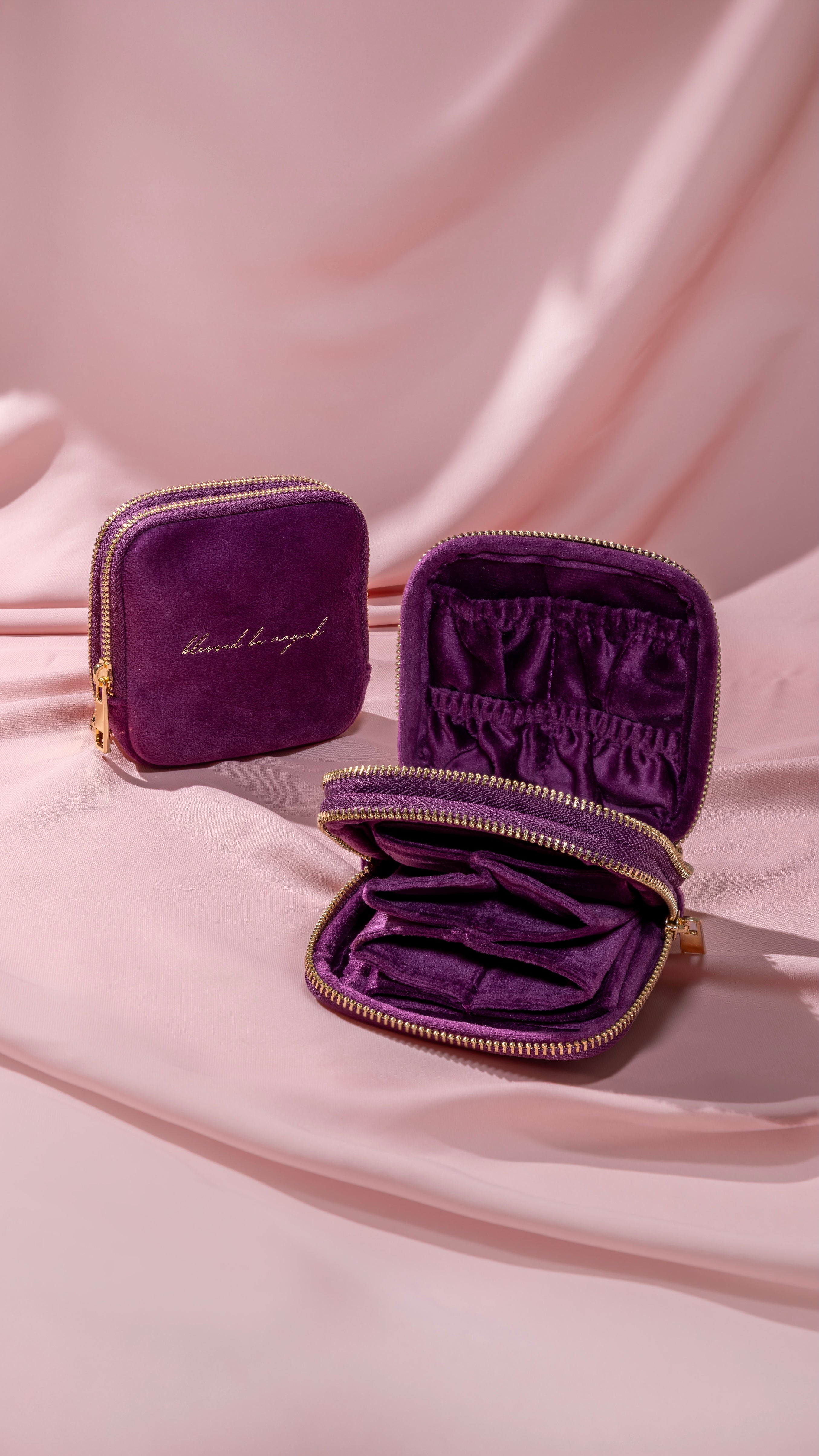 Two small purple velvet zippered cosmetic pouches on draped pink fabric, one closed with gold script lettering and one open showing interior compartments.