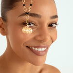 A close-up of a woman wearing a gold chain necklace with black beads and a gold pumpkin-shaped charm, set against a light blue background.