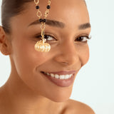 A close-up of a woman wearing a gold chain necklace with black beads and a gold pumpkin-shaped charm, set against a light blue background.