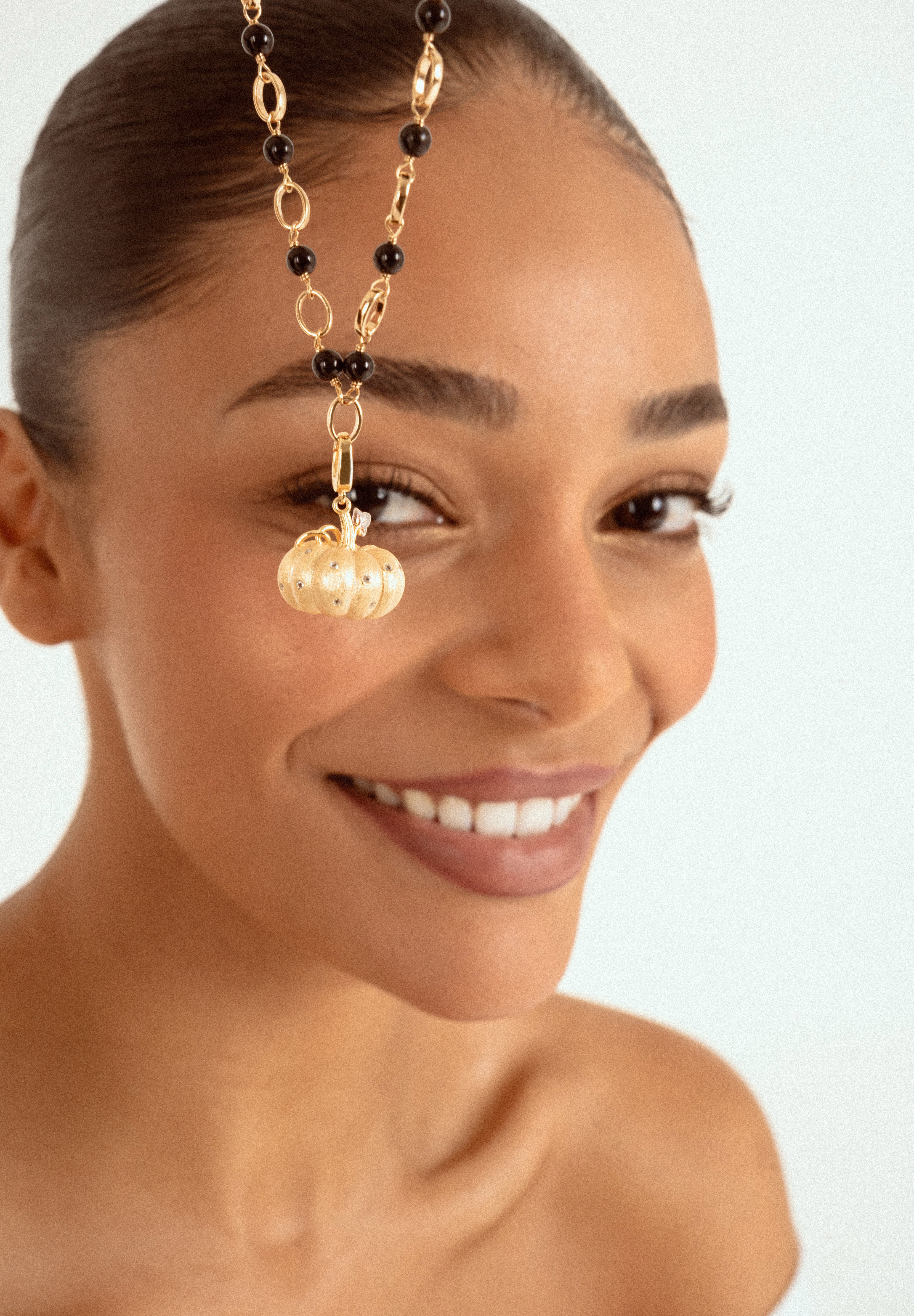 A close-up of a woman wearing a gold chain necklace with black beads and a gold pumpkin-shaped charm, set against a light blue background.