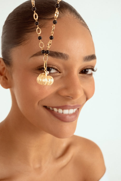 A close-up of a woman wearing a gold chain necklace with black beads and a gold pumpkin-shaped charm, set against a light blue background.