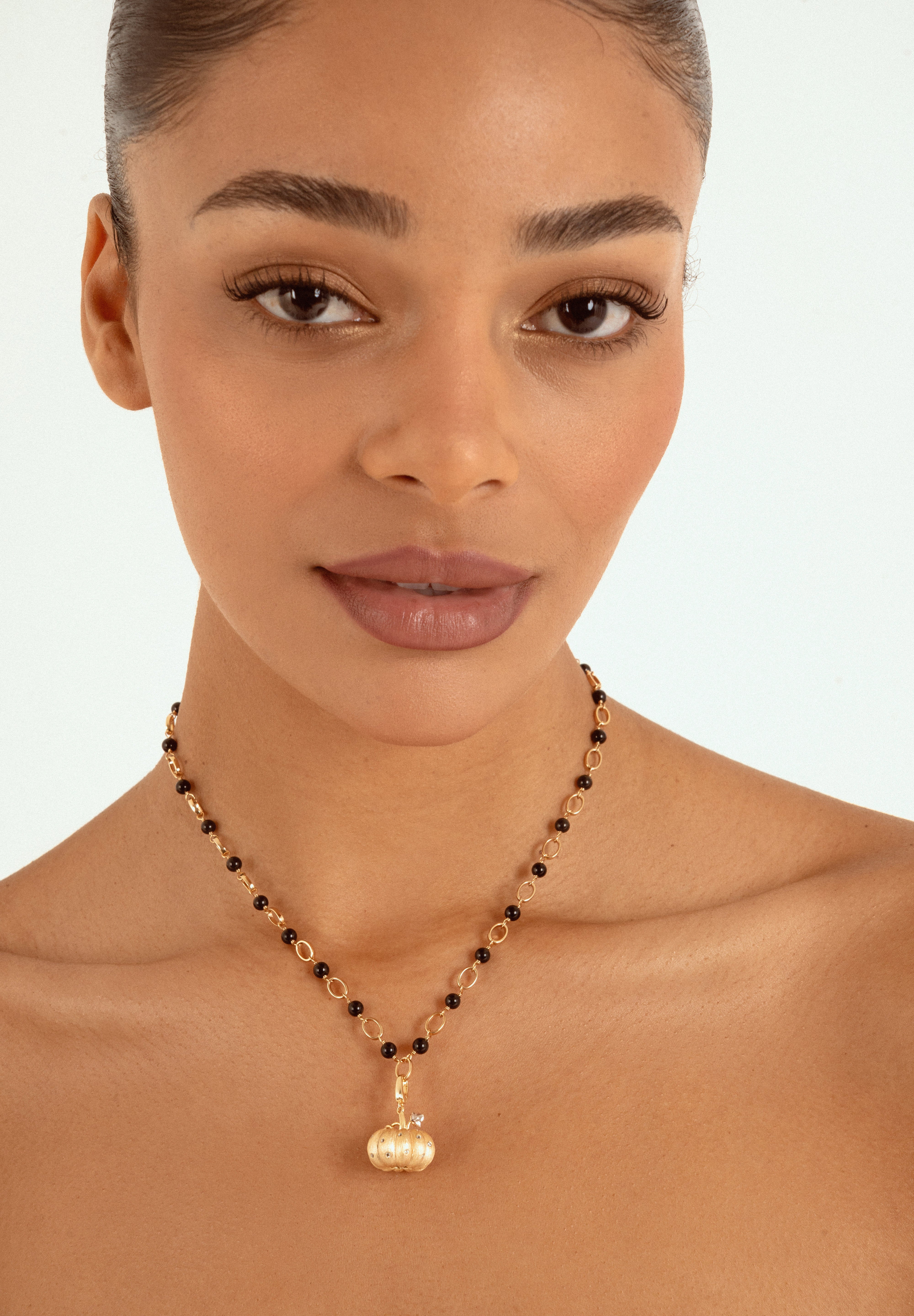 A close-up of a woman wearing a delicate gold necklace with small black beads and a gold pumpkin-shaped pendant, set against a clean white background.