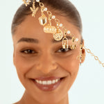 A smiling woman with her hair pulled back holds up s a delicate gold chain headpiece adorned with small charms, including sun, pumpkin, acorn, butterly, acorn charms and pearl-like beads, against a light blue background.