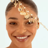 A smiling woman with her hair pulled back holds up s a delicate gold chain headpiece adorned with small charms, including sun, pumpkin, acorn, butterly, acorn charms and pearl-like beads, against a light blue background.