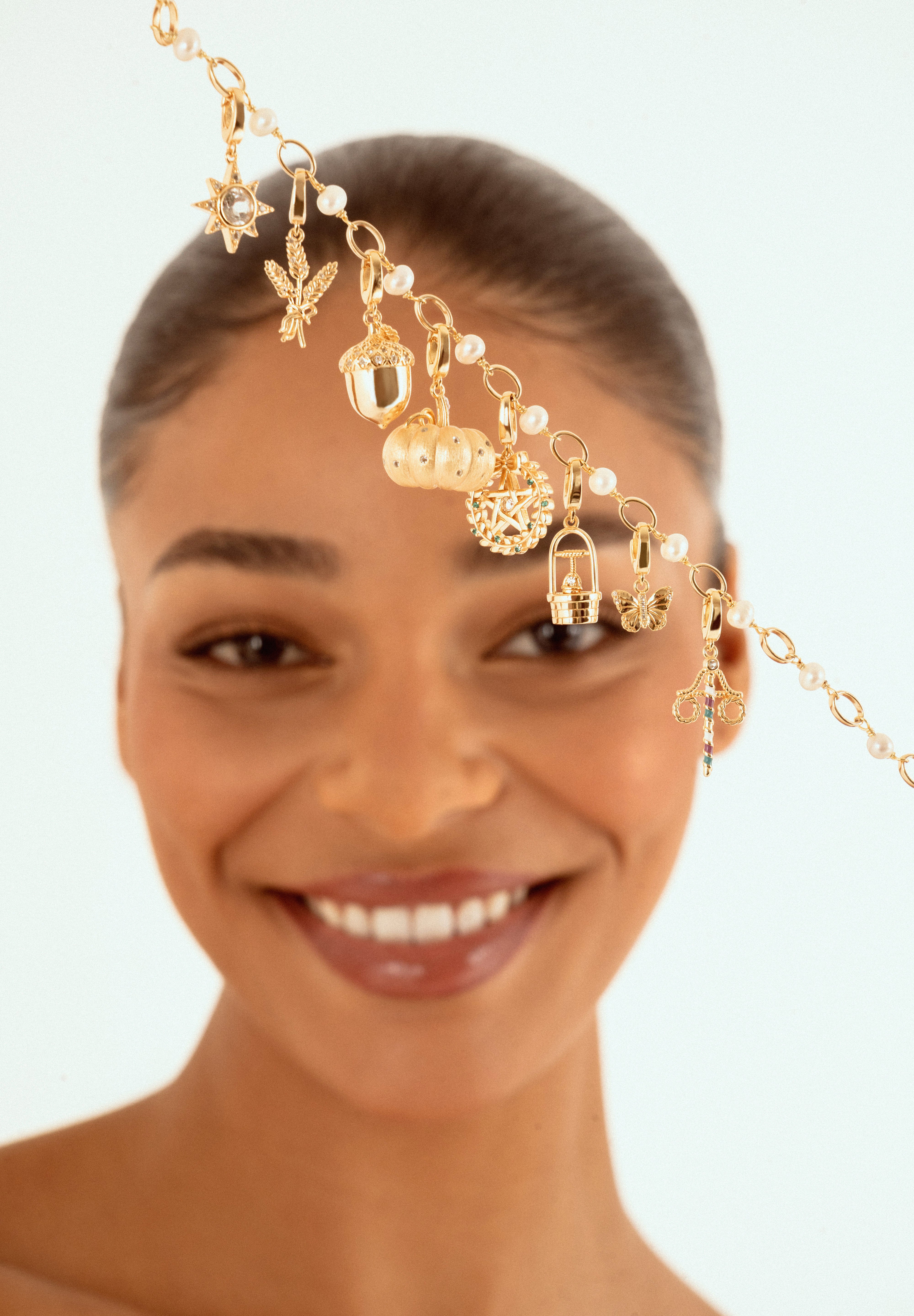 A smiling woman with her hair pulled back holds up s a delicate gold chain headpiece adorned with small charms, including sun, pumpkin, acorn, butterly, acorn charms and pearl-like beads, against a light blue background.