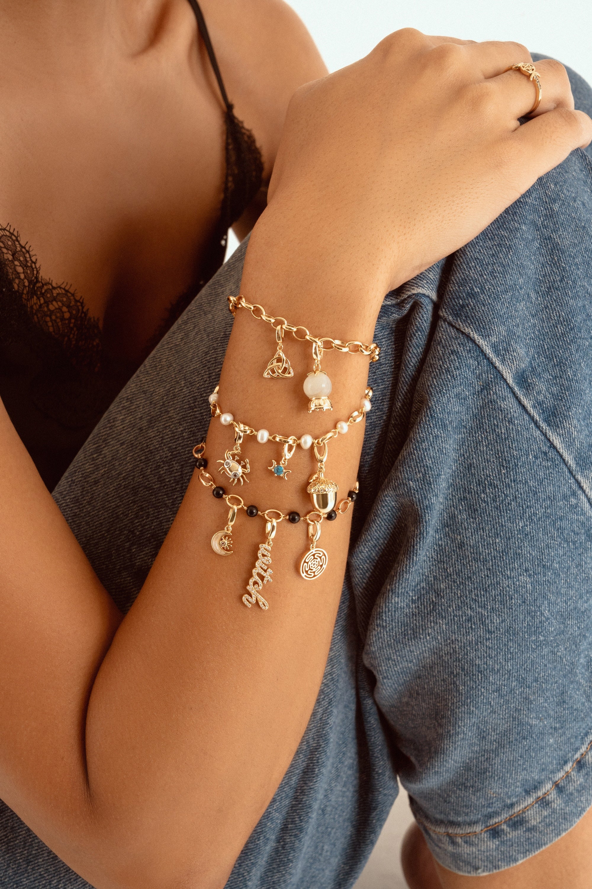 A close-up of a person wearing a stack of three delicate charm bracelets on their wrist, paired with a black lace camisole and blue denim shorts.