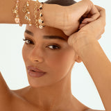 A close-up of a woman's face and raised arms, showcasing delicate gold-toned charm bracelets with rose quartz and pearl beads on her wrist against a clean white background.
