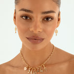 A close-up of a woman wearing a delicate gold charm necklace and matching drop earrings against a clean white background.