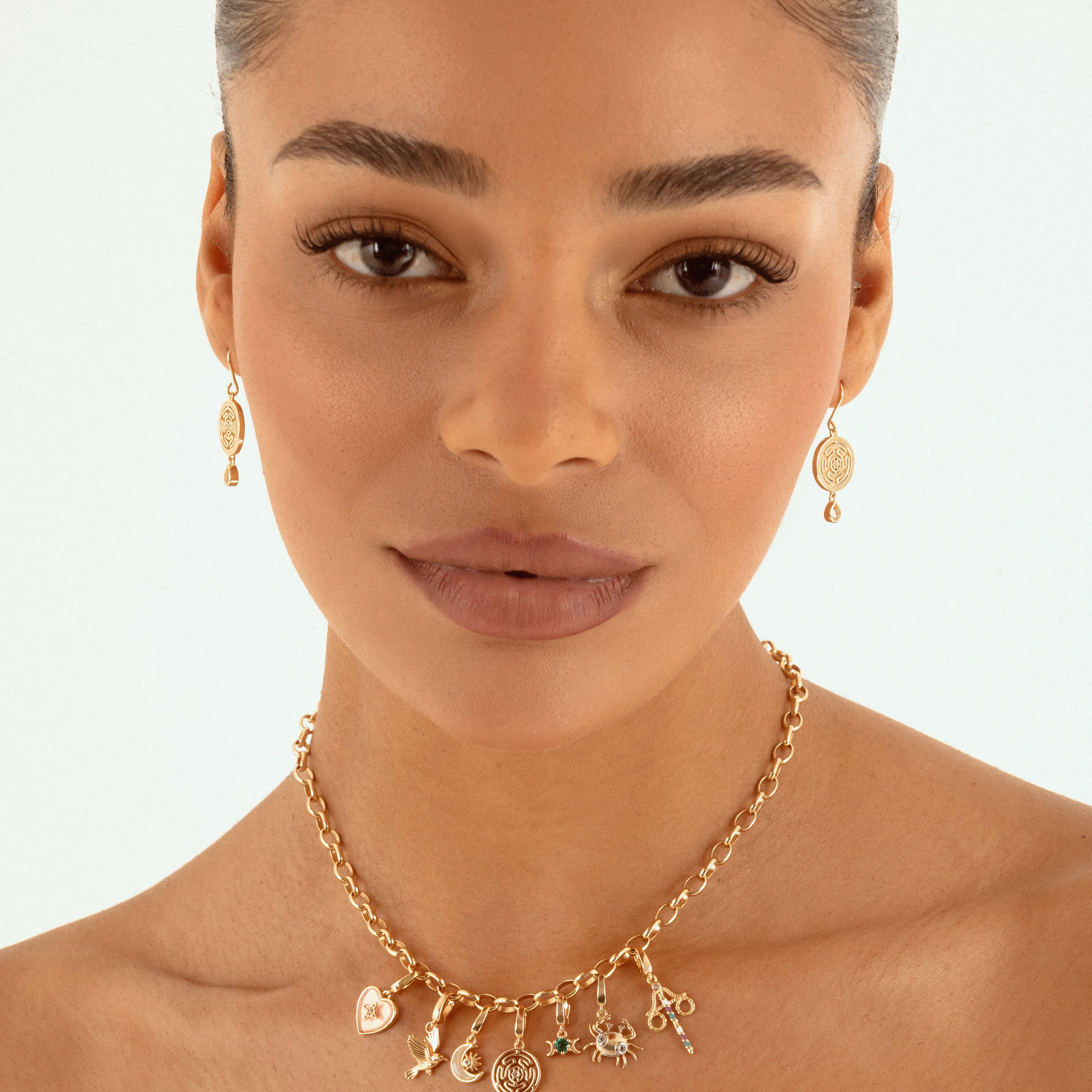 A close-up of a woman wearing a delicate gold charm necklace and matching drop earrings against a clean white background.
