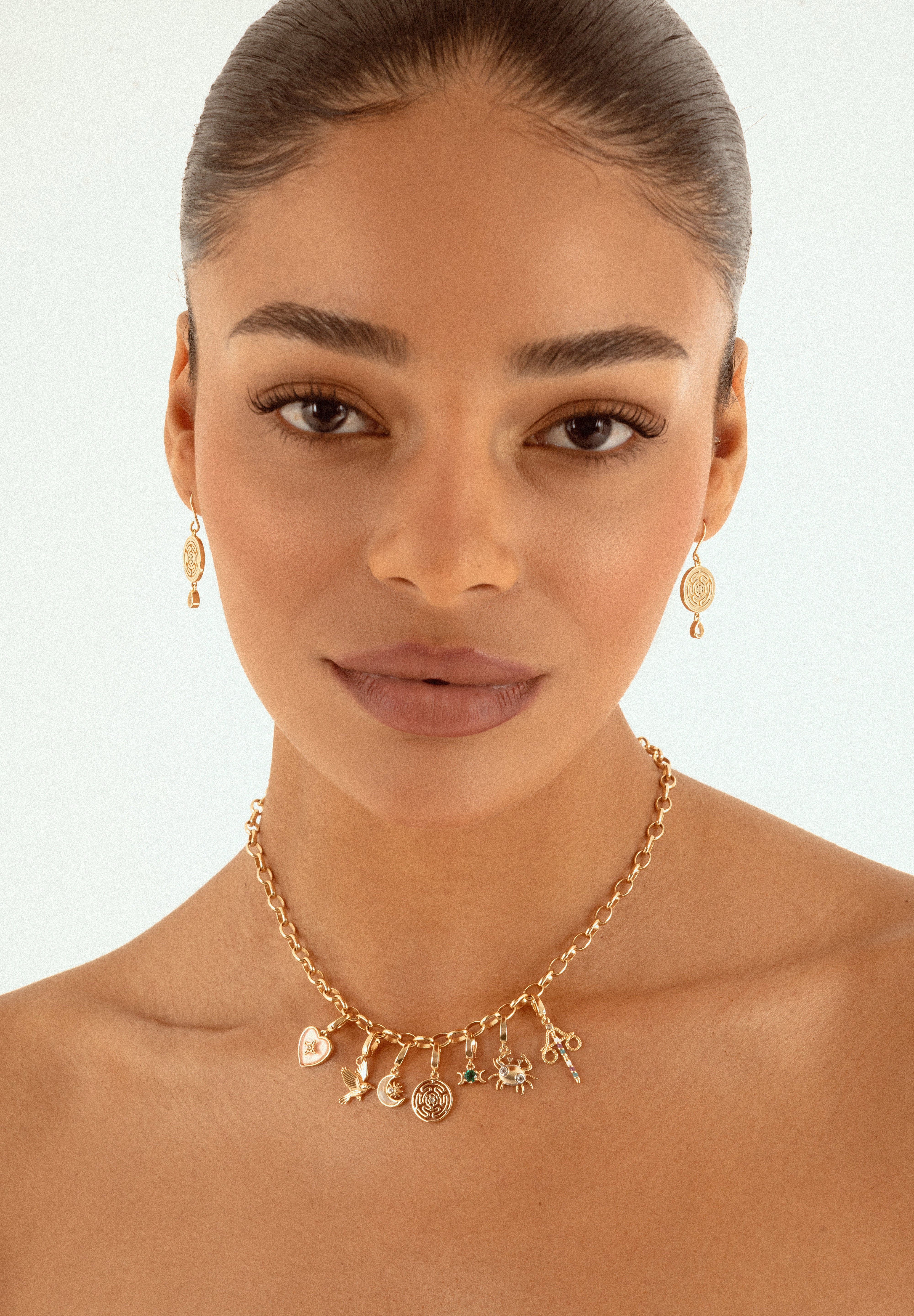 A close-up of a woman wearing a delicate gold charm necklace and matching drop earrings against a clean white background.