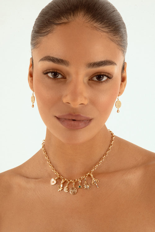 A close-up of a woman wearing a delicate gold charm necklace and matching drop earrings against a clean white background.