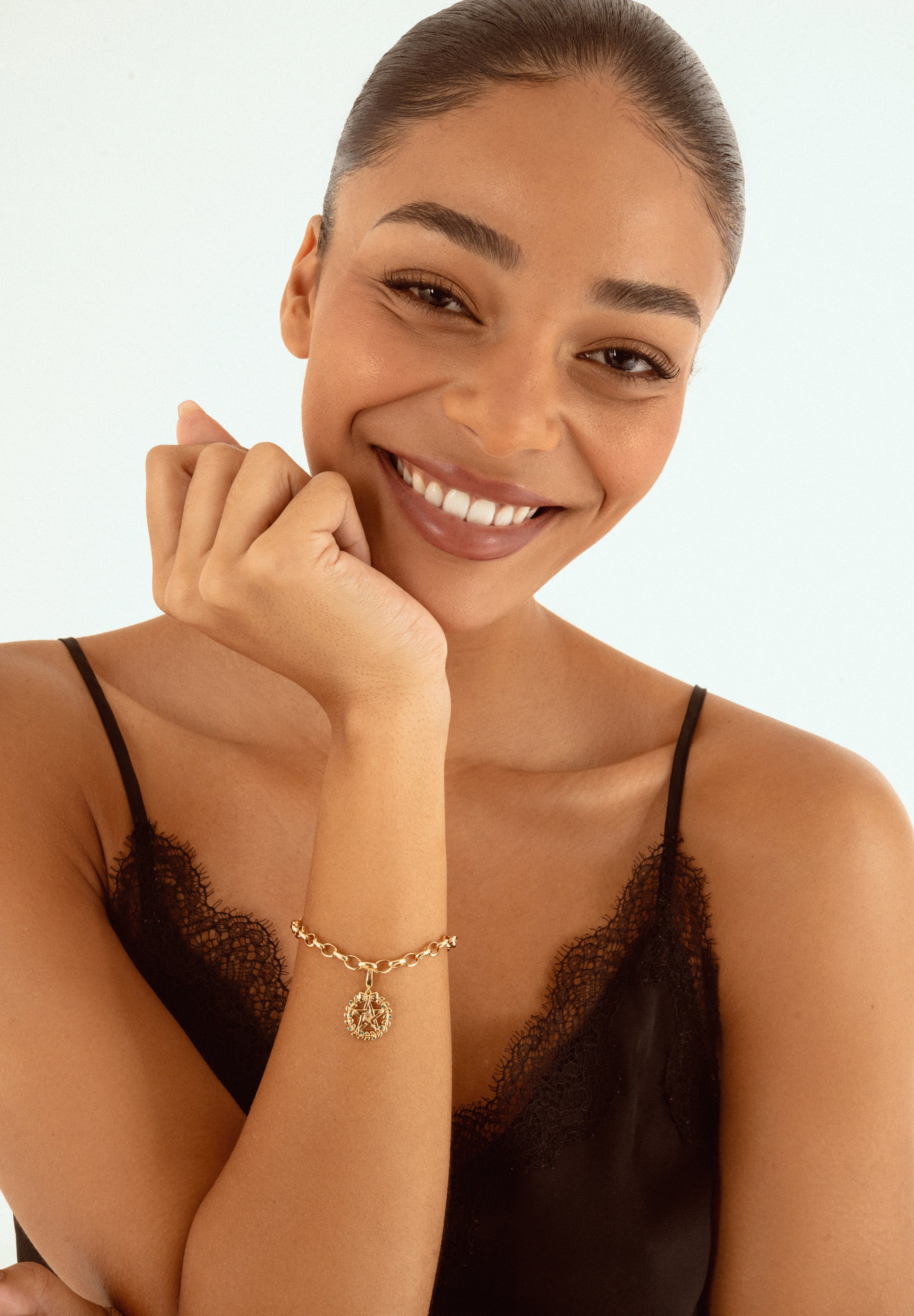 A smiling woman with her hair pulled back wears a delicate gold chain bracelet with a Pentacle sign charm, paired with a black lace camisole top against a clean white background.
