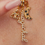 Close-up of a person running her fingers through a gold necklace with three charms: a triquetra symbol, a script ‘magickal’ charm, and a gold crescent moon with black and white stones.