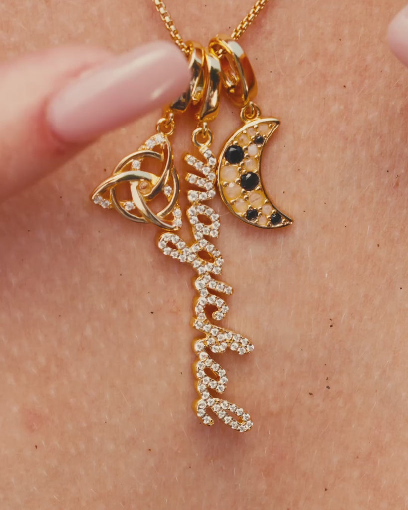 Close-up of a person running her fingers through a gold necklace with three charms: a triquetra symbol, a script ‘magickal’ charm, and a gold crescent moon with black and white stones.